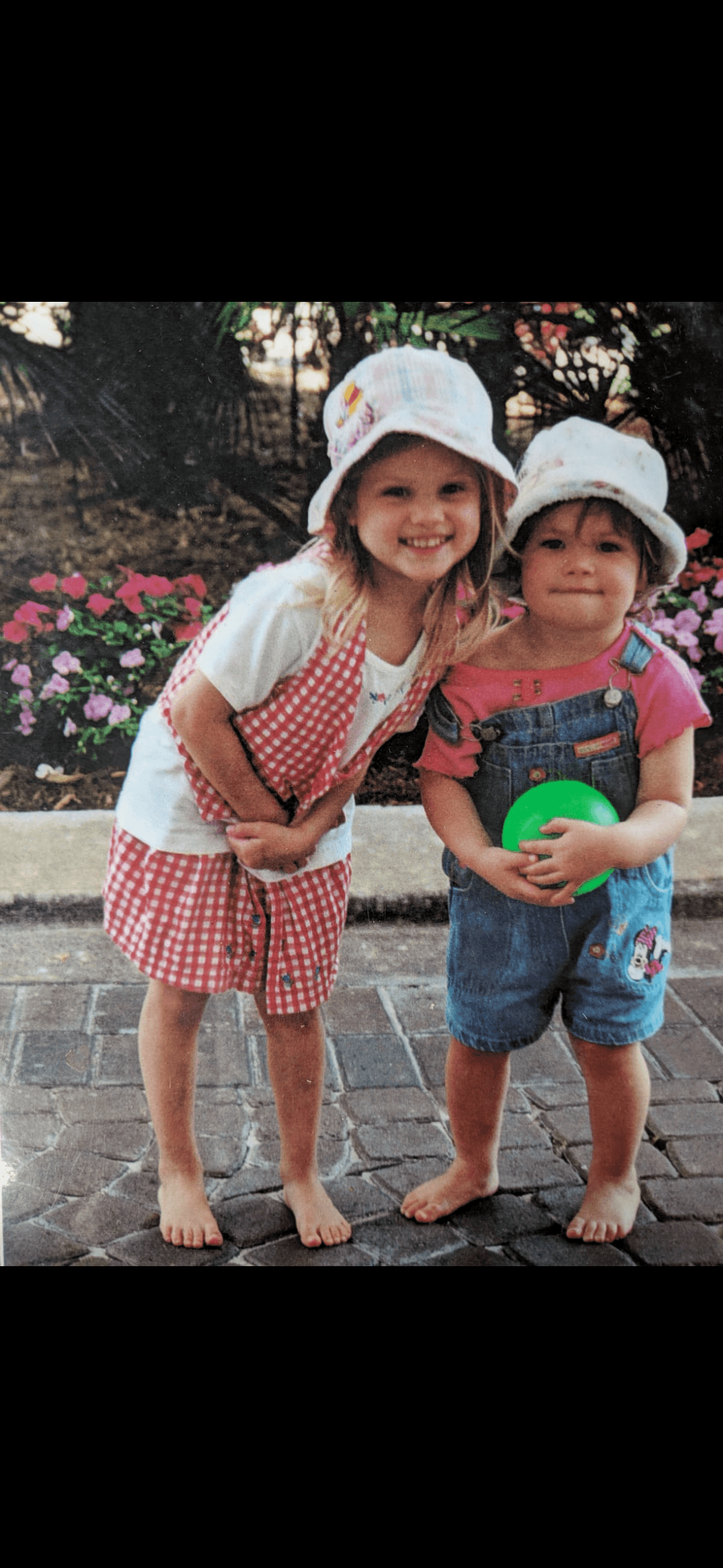 Ellie and her sister as little kids, both in bucket hats, standing in front of flowers.